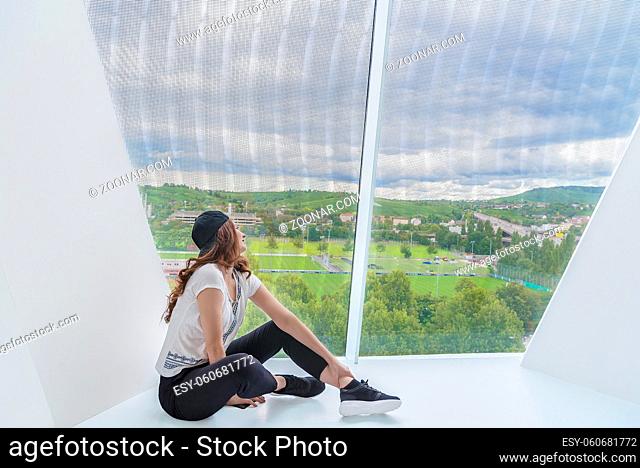 Young attractive woman, dressed in casual clothing, sits on a white floor from a modern building and looks through a very tall window