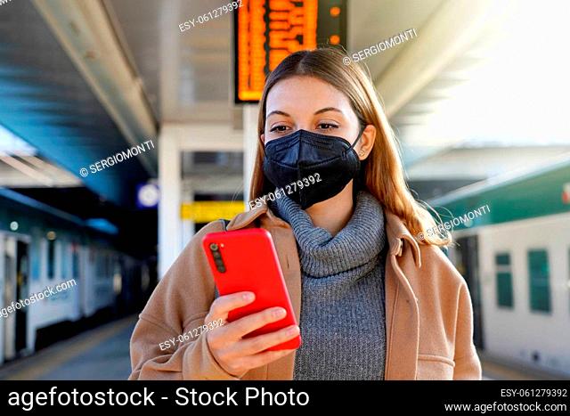 Young woman with black medical mask KN95 FFP2 using telephone in the train station with timetables on the background