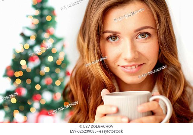 holidays, drink, winter and people concept - close up of happy young woman with cup of coffee or tea over christmas tree lights background