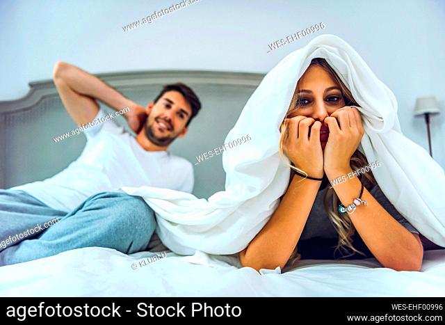 Portrait of young woman lying in bed at home with boyfriend in background