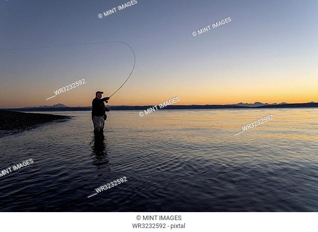 A silhouette view of a fly fisherman casting for salmon and searun coastal cutthroat trout from a salt water beach at a beach on the north west coastline of the...