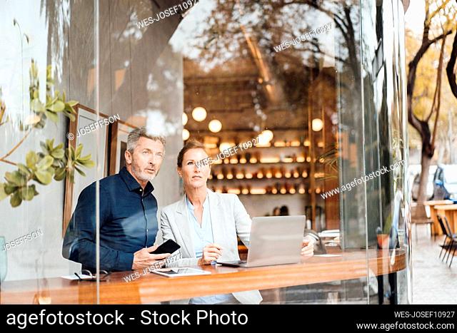 Businessman holding mobile phone standing by businesswoman in cafe seen through window