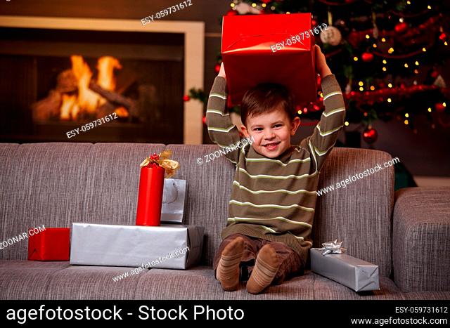 Portrait of happy little boy sitting on couch holding with christmas presents, holding box overhead