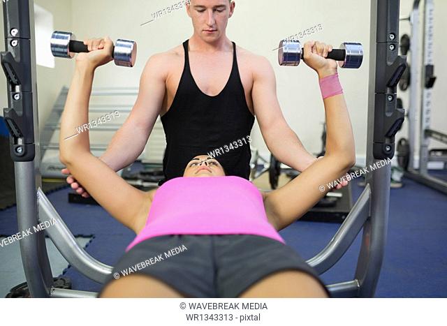 Serious instructor helping lying woman exercising with dumbbells