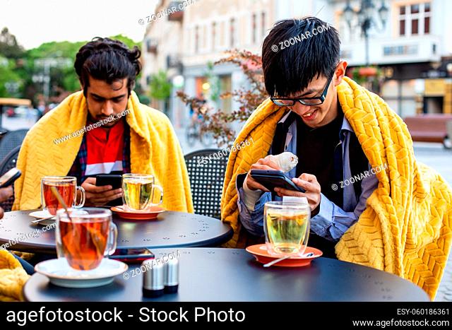 Three close diverse friends at the outdoor cafe, sitting covered with yellow plaids, watching their smartphones. Young men ordering food using online menu from...