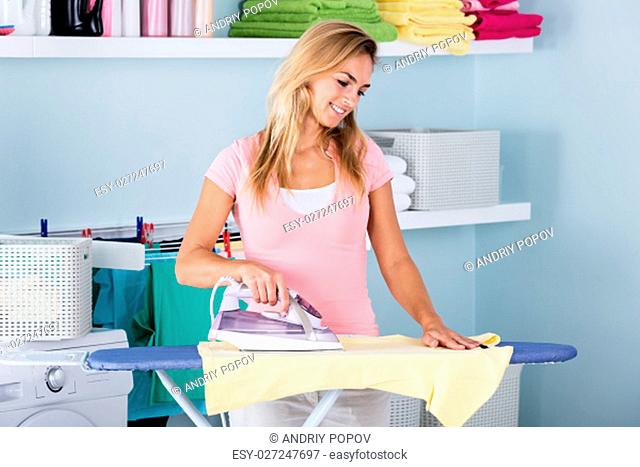 Smiling Woman Ironing Clothes Using Iron On Ironing Board After Laundry At Home