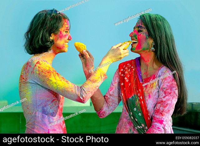 TWO YOUNG WOMEN GIVING EACH OTHER SWEETS WHILE CELEBRATING HOLI
