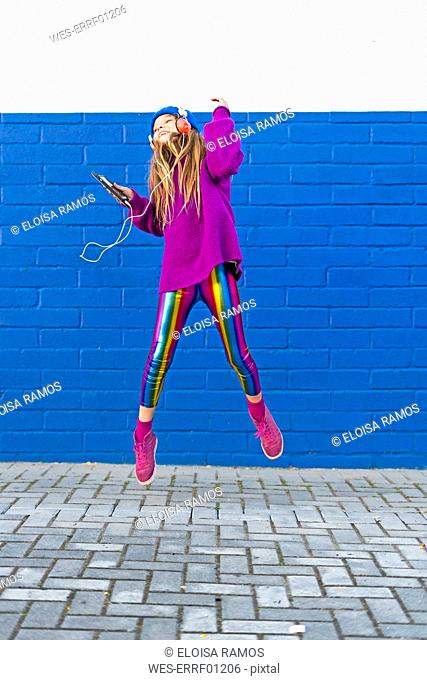 Happy girl with headphones and smartphone jumping in the air in front of blue wall