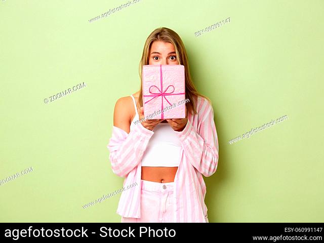 Image of shy cute girl giving you a present, hiding behind gift box and peeking at camera, standing over green background