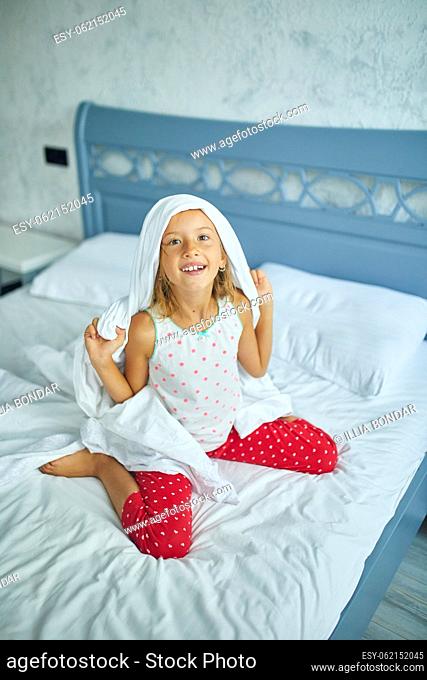 Happy little girl wakes up, joyful, blanket on the head, from sleep on a big and cozy bed white linen in the at home, family lifestyle, comfortable pillow, bed