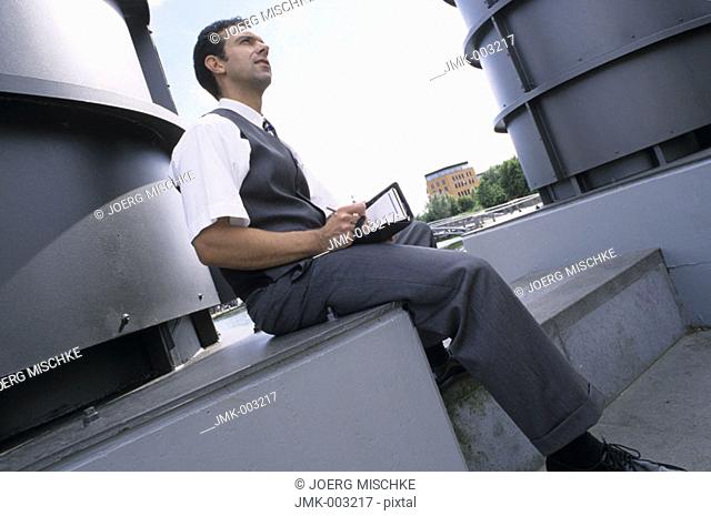 A young man, businessman, 25-30 30-35 35-40 years old, sitting outside in an industrial estate with a calendar, datebook