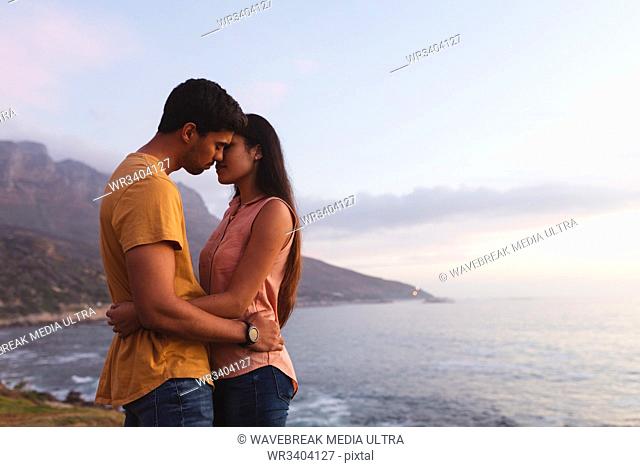 Side view of a young mixed race couple standing on a beach embracing and about to kiss at sundown