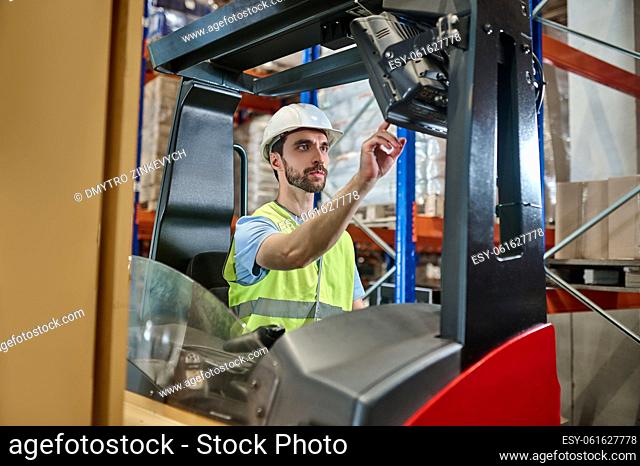 Serious concentrated young storehouse worker in a helmet and reflective vest starting an electric forklift