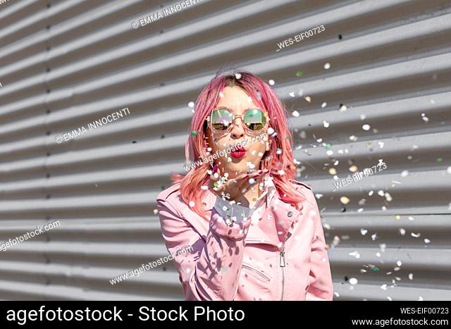 Young woman with pink hair blowing paper confetti in front of corrugated wall