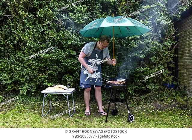 A Young Man Cooks Food On A Barbeque During A Rain Shower, Lewes, East Sussex, UK