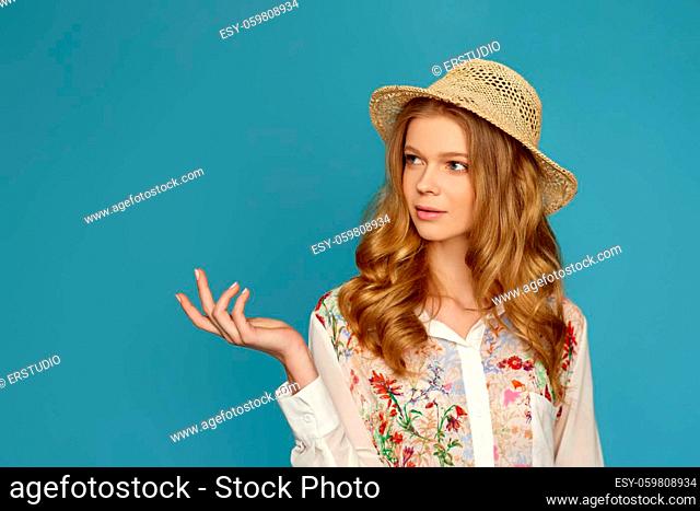 portrait of beautiful young blonde woman in straw hat pointing to the side on blue background