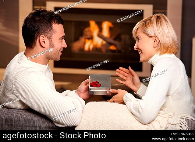 Young romantic couple sitting on couch in front of fireplace at home, man giving gift, side view
