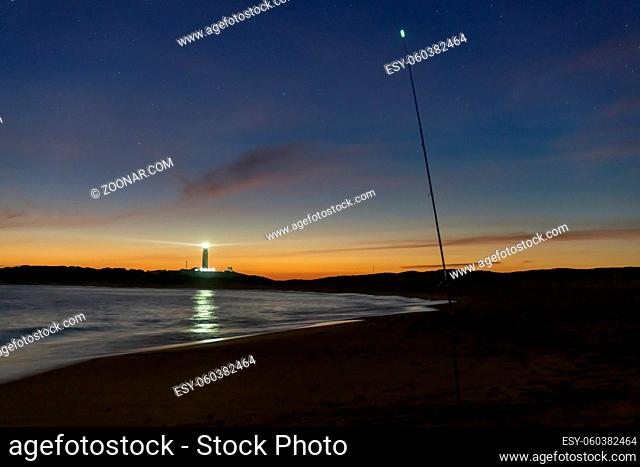 A fishing rod with luminescent tip on a beach at night with lighthouse behind