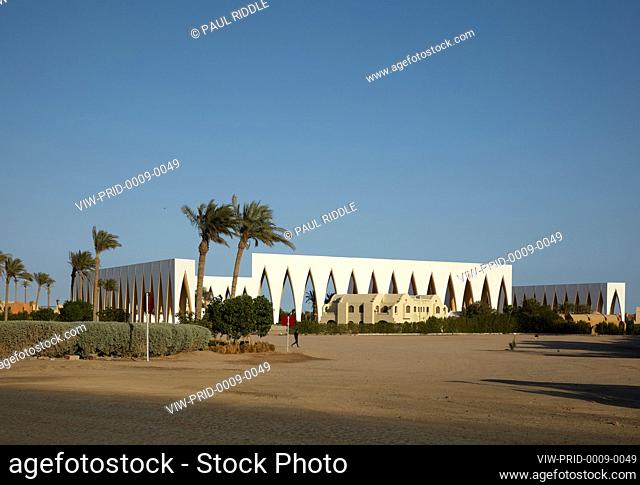 Oblique contextual view with sand foreground. Man walking middle frame. Gouna Festival Plaza, El Gouna, Egypt. Architect: Studio Seilern Architects, 2021