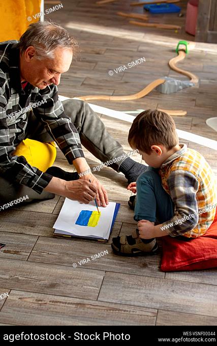 Grandson with grandfather drawing Ukrainian flag on paper at home