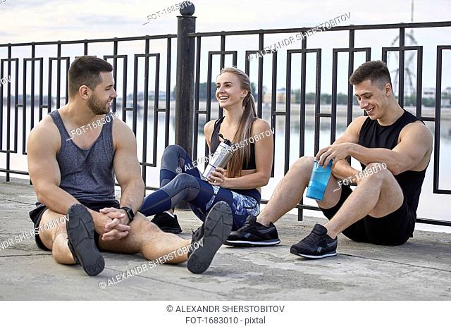 Smiling young athletes taking break while sitting on footpath by railing