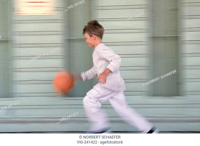 portrait, full-figure, 8 years old boy wearing grey trouser and pullover passes with an orange basketball by a grey-blue painted fassade  - GERMANY, 09/04/2004