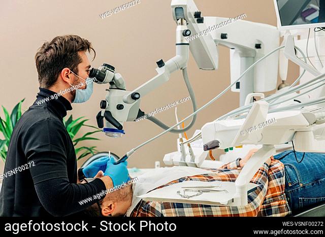 Young dentist examining patient's teeth through microscope in clinic
