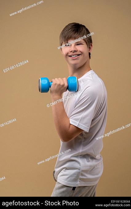 Smiling cheerful teenage boy holding the dumbbell in his hand and looking into the distance