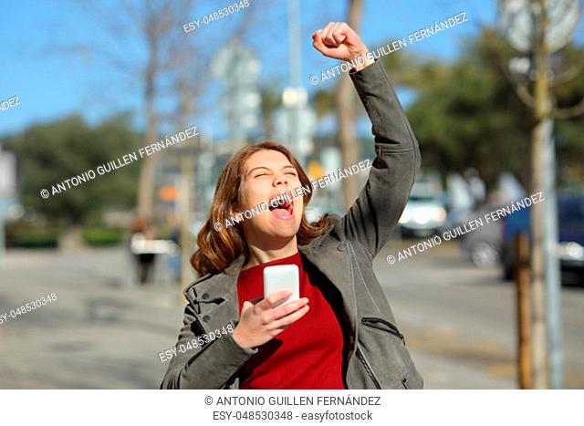 Excited teen celebrating online good news holding a smart phone in the street