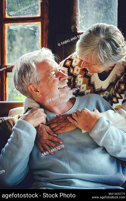 Happy senior couple hug and enjoy time together at home sitting on che chair and smiling each other. Concept of old people mature relationship
