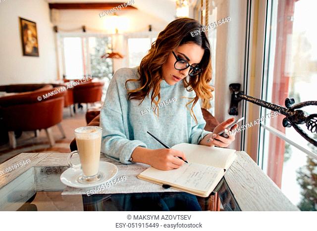 Young business woman in cafe writes notes in notebook from smartphone