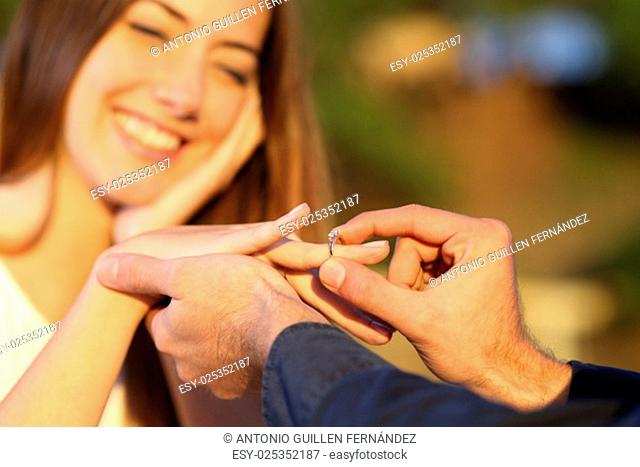 Boyfriend putting a engagement ring in his girlfriend finger outdoors with the smile of the girl in the background