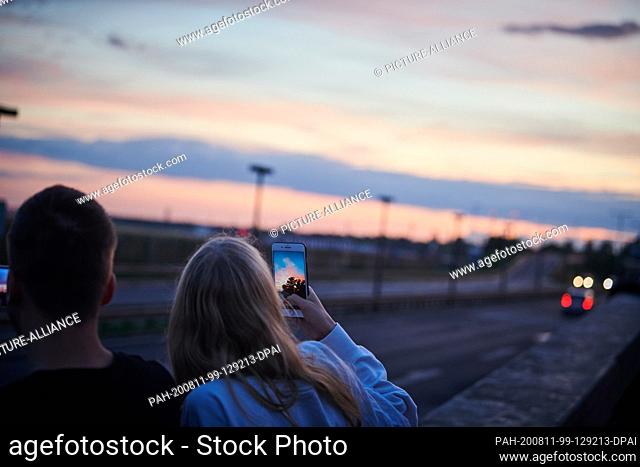 26 July 2020, Berlin: Planespotter on the motorway at Tegel Airport. A young couple is waiting at dusk to see landing planes