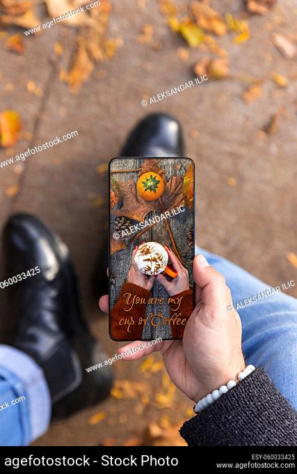 Man received love photo message on smartphone while sitting outdoors in a park full of autumn leaves on the ground. Coffee time