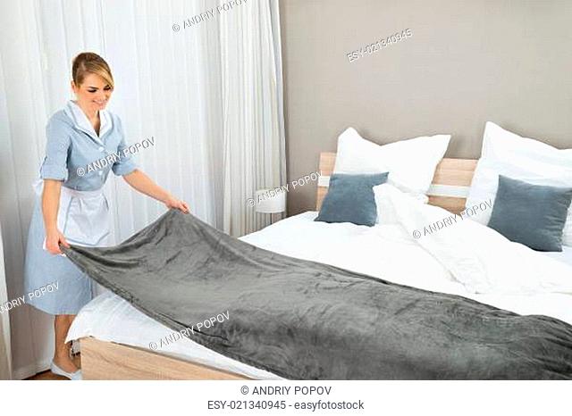 Happy Female Housekeeping Worker Making Bed In Hotel Room