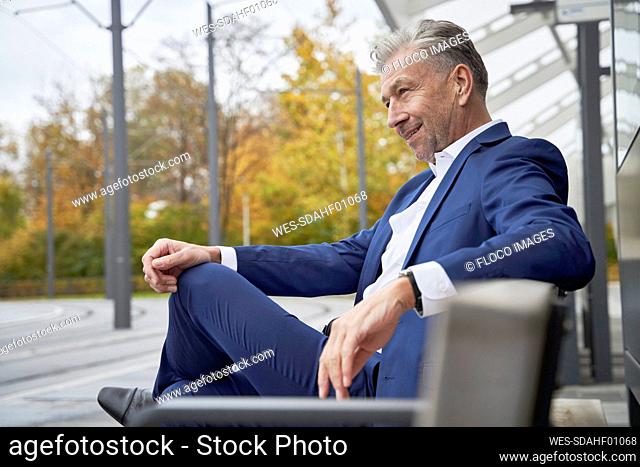 Smiling confident senior male professional sitting on bench while waiting at bus stop