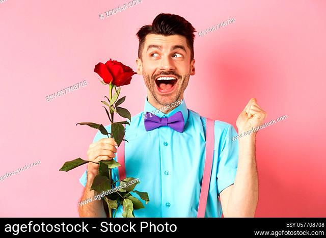 Romance and Valentines day concept. Cheerful man in bow-tie screaming from happiness, holding red rose and jumping on date, standing over pink background