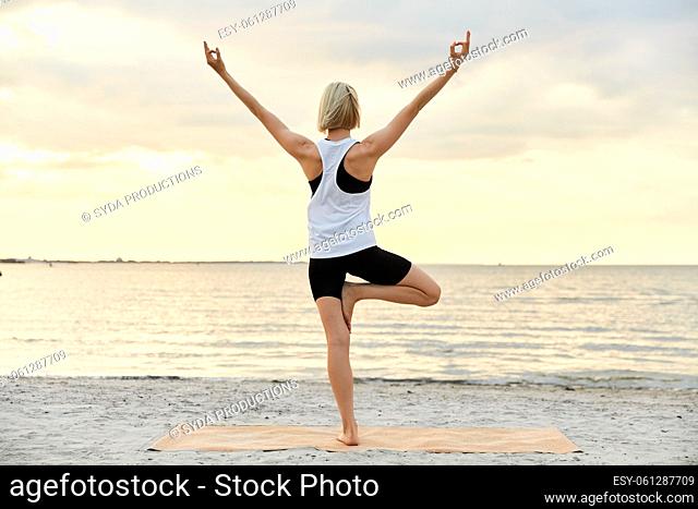woman doing yoga tree pose on beach over sunset