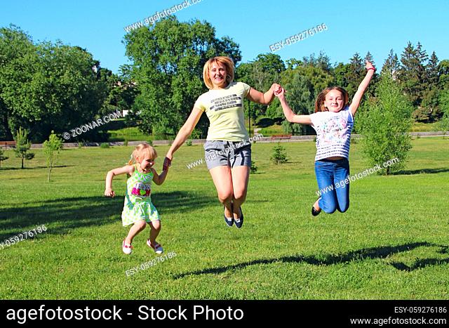 Happy mother having fun jumping with her daughters on green grass. Happy family team. Family enjoying summer vacations. People enjoying family holiday