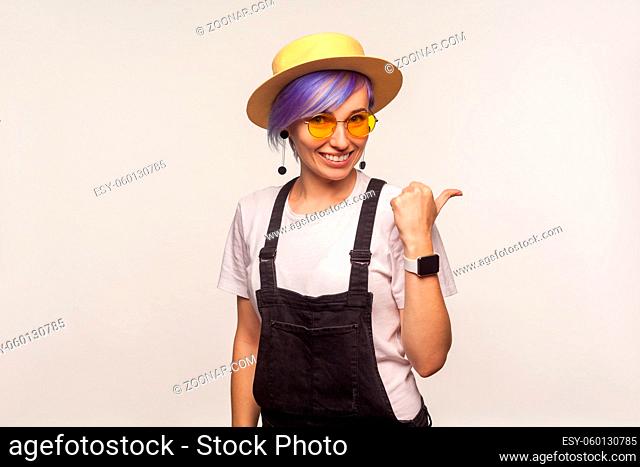 Portrait of modern glamour hipster girl with violet short hair in sunglasses and hat smiling and pointing to the side, copy space for advertisement