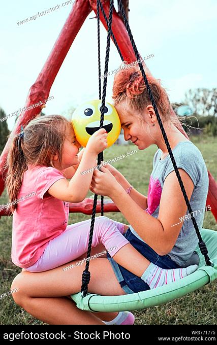 Teenage girl playing with her younger sister in a home playground in a backyard. Happy smiling sisters having fun on a swing together on summer day