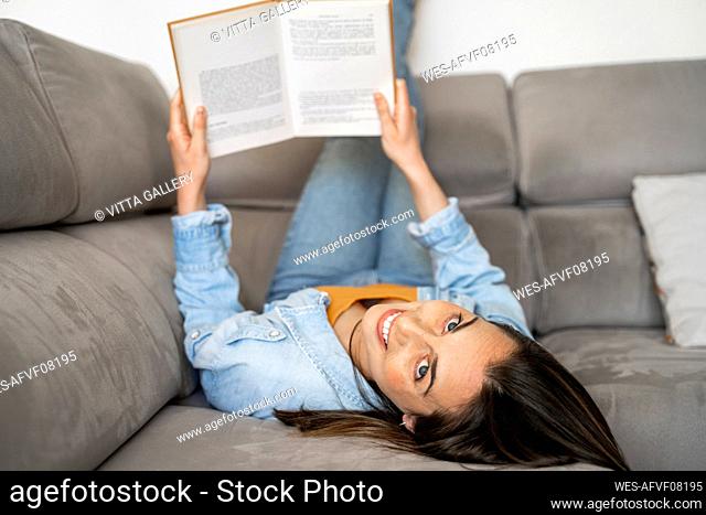 Smiling woman with feet up holding book while lying on sofa at home