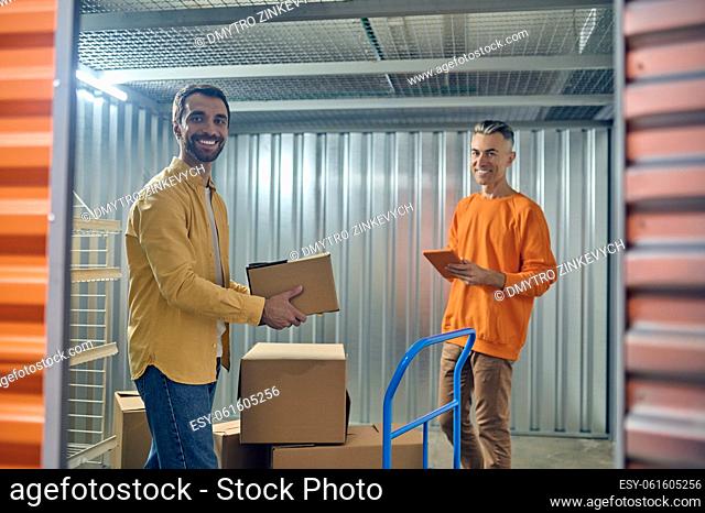 Warehouse worker with the cardboard box and his colleague with the tablet computer standing in the cargo container