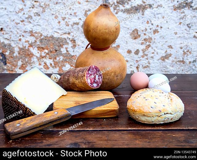 An Iberian pork sausage on an old wooden board with an antique knife, a loaf of Spanish rustic seed bread, cheese, three multicolor eggs and a bottle gourd