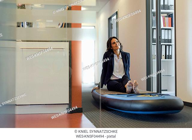 Businesswoman sitting on paddle board, daydreaming in office