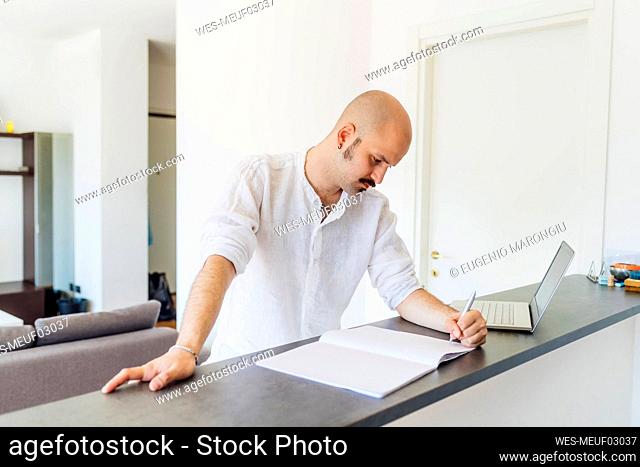 Mustache man writing on book by laptop at home