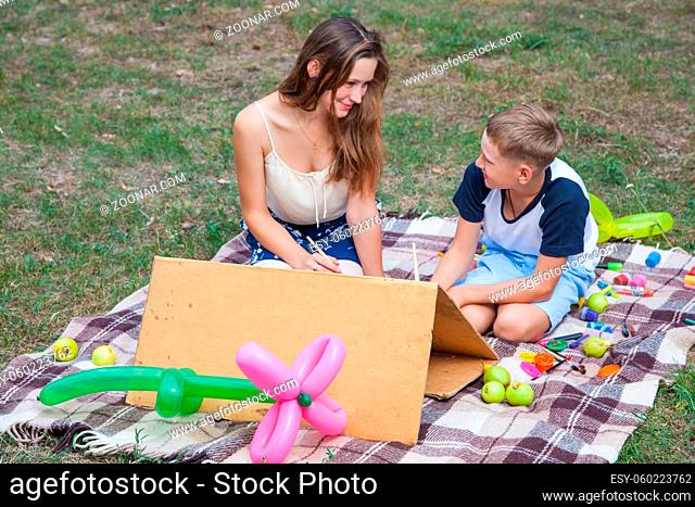 Older sister trying to teach her brother to paint and posing in the park on summer time, looking and smiling