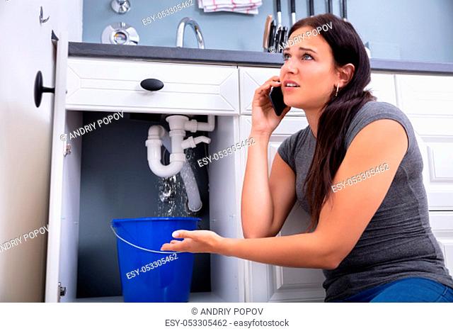 Worried Woman Calling Plumber While Collecting Water Leaking From Sink Using Utensil