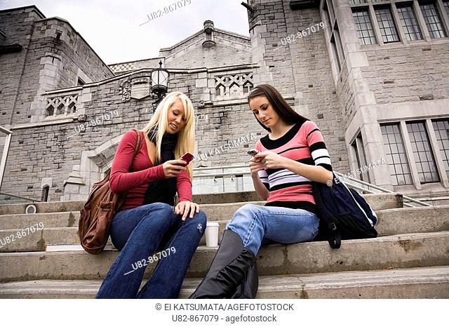 Two female college students sitting on the steps of a building texting on the cell phones, University of British Columbia, Vancouver, BC, Canada