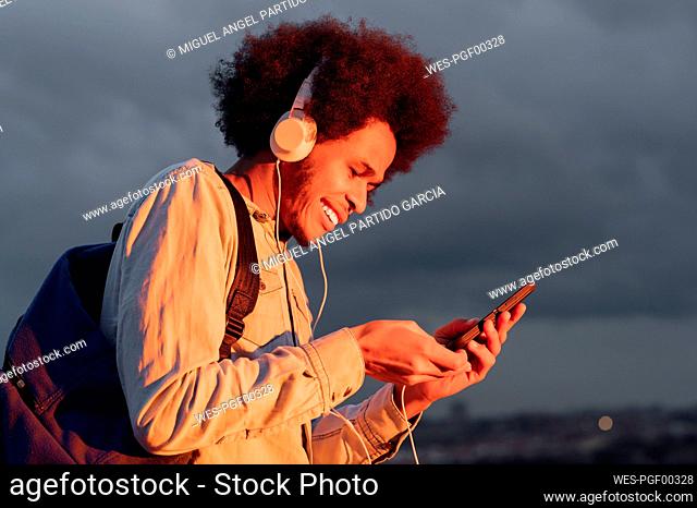 Smiling man listening music and using smart phone against cloudy sky at sunset